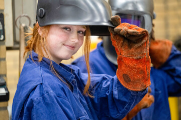 A young female welder in a blue coverall lifts her welding helmet with a gloved hand, smiling at the camera in an industrial setting.