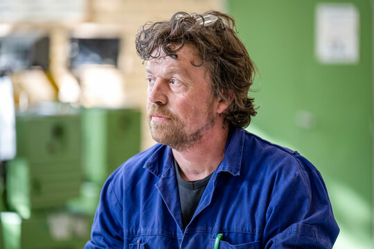 A man with curly hair wearing a blue work uniform is looking to the side with a reflective expression, indoors, likely in a workshop environment. - Powered by Adobe