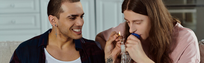 Obraz premium A gay couple, dressed casually, laughing joyfully together at home, lighting marijuana in the glass bong