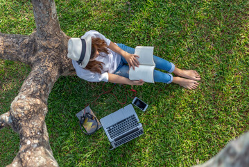Girl enjoy listening music and reading a book and play laptop on the grass field of the park in the...