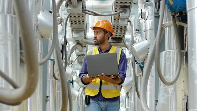 Young asian male technical foreman in safety uniform inspects maintenance work holding a laptop to look at plumbing and electrical systems on the roof of a building, Industrial background