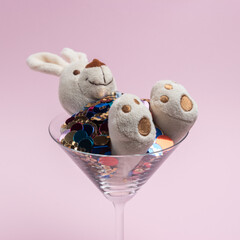 Bunny toy in martini glass full of colorful glitters on pink background. Creative minimal concept.