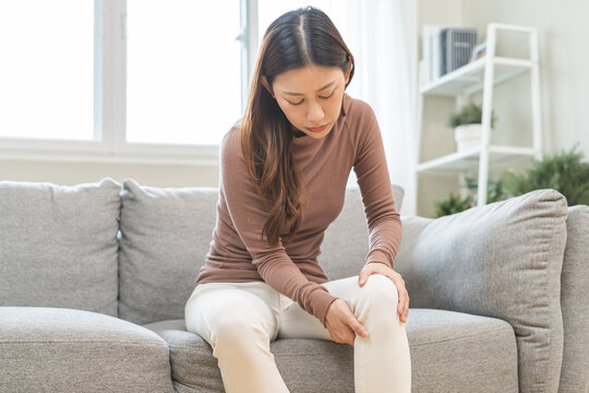 Health care of knee pain concept, suffering asian young woman arthritis sitting joint ache, sore cramp or sprain tendon in leg on sofa at home. Osteoarthritis chronic disease of body problem, recovery