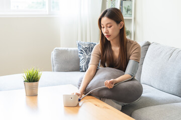 Hypotension, asian young woman sitting on couch checking blood high pressure and heart rate with digital pressure monitor machine making self check up on her arm at home. Healthcare and medical.
