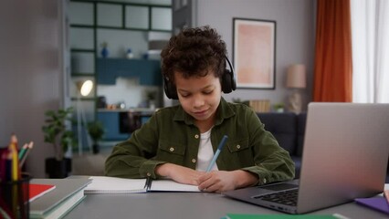 African American multiracial little boy in headphones listening online teacher son kid child pupil at home education studying learning computer distant class lesson laptop video call writing task