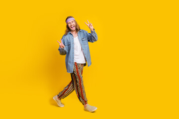 Full body photo of attractive young hippie man walking show v-sign wear trendy denim outfit isolated on yellow color background