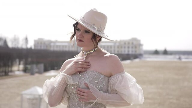 Elegant woman in stylish hat and gown in open field, surrounded by historic buildings in sunlight