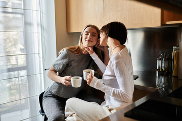 Two women in a hotel kitchen, sipping coffee together, showcasing love and unity.