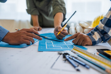 An architect engineer meeting is taking place at a desk covered with blueprints. They are...