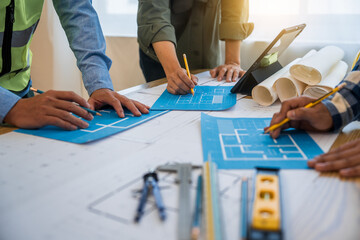 An architect engineer meeting is taking place at a desk covered with blueprints. They are...