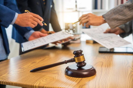 A team meeting of business people and a lawyer in formal suits is taking place at a desk, discussing a contract and various aspects of the law and litigation.