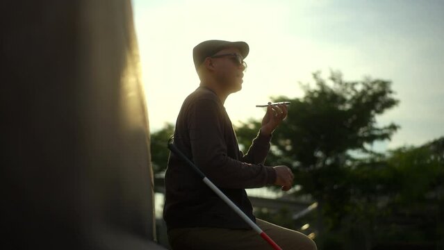 The young blind man using voice control app on smartphone while sitting on a bench against beautiful sunlight in the city park, disabled visually impaired man talking on cellphone for communication.
