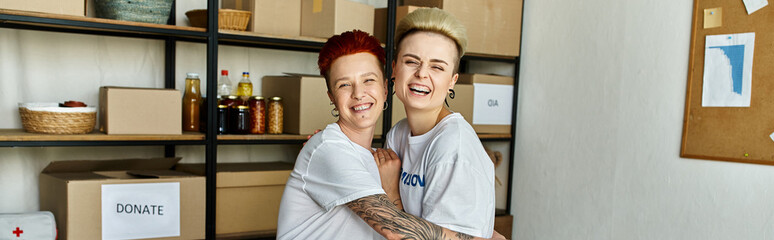 A young lesbian couple wearing volunteer t-shirts stands together in a room, working on a...