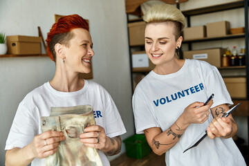Two women, a young lesbian couple, stand side by side in volunteer t-shirts, united in charity work.