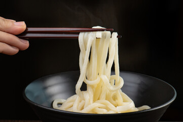 A close up of a person holding chopsticks with udon noodles above a black bowl.