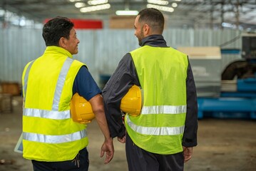 Two male factory workers wearing yellow safety vests walk and talk in a large industrial building.