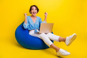 Full length photo of overjoyed charming girl dressed print blouse sit on pouf with laptop win bet isolated on yellow color background