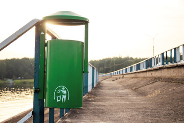 green color recycling bins in a public park near the river. Metal trash bin in the park with blurry sunny background and forest. Bridge and dam with street, bicycle and pedestrian path