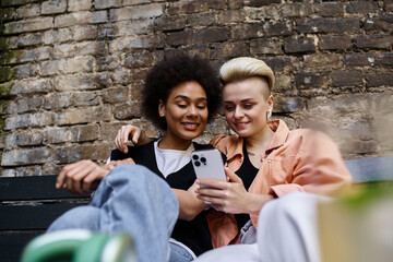 Two women, a diverse and beautiful lesbian couple, sit on a bench in a cafe, engrossed in their shared cell phone.