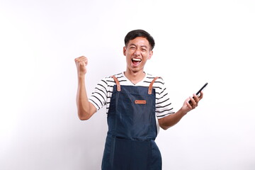 Young asian man barista bartender barman employee in blue apron white t-shirt work in coffee shop using mobile cell phone do winner gesture isolated on white background. Small business startup concept