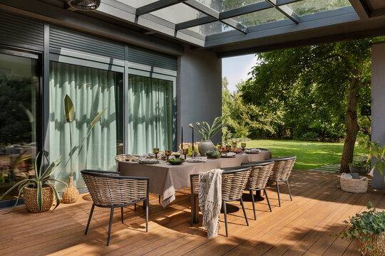 Beautiful composition of cozy terrace with big table, garden chair, plaid, plants in flowerpots, tableware, fruits in bowl, buns, brown tablecloth and personal accessories. Home decor. Template.