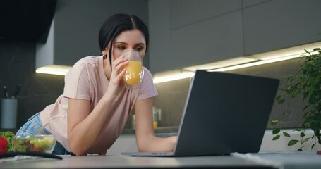 Smiling woman working on laptop which standing on kitchen table and drinking orange juice. - Powered by Adobe