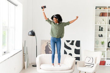 African-American woman celebrates with arms wide open, standing on a sofa in a bright, modern room,...