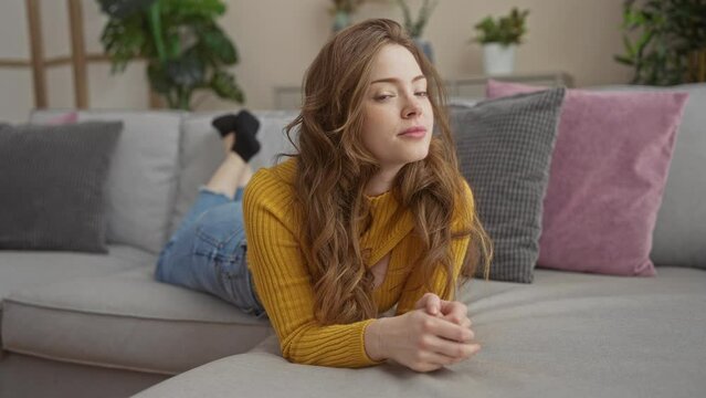 A blonde, young and beautiful woman relaxes indoors at home, laying on a grey couch in a living room surrounded by cushions and plants.