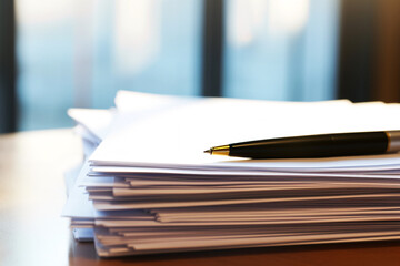 Fototapeta premium Pile of documents on a desk with a black pen placed on top bathed in warm sunlight