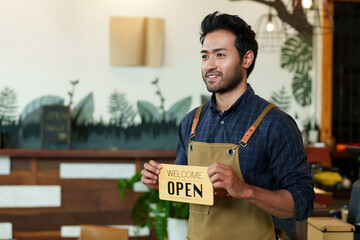 Multiethnic male cafe owner businessman standing smiling and holding a sign saying OPEN. To promote...