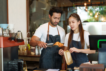 Couple Asian baristas in aprons, engaging over pastry packaging, with professional coffee equipment...
