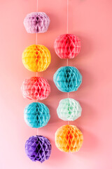 Colorful paper honeycomb balls in shades of purple, yellow, pink, turquoise, and mint green hang vertically against a pink background, creating a vibrant and festive display