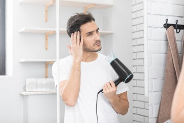 Young handsome man standing in front of the mirror in the bathroom and holding hair dryer. Morning...