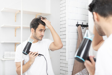 Young handsome man standing in front of the mirror in the bathroom and holding hair dryer. Morning...