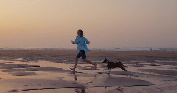 Cinematic commercial shot of woman run wild, young and free on sandy beach during low tide in sunset light. Happy and excited, concept of freedom and happiness. Running happily with pet dog