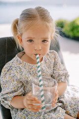 Little girl drinks water from a glass through a striped straw while sitting in a chair