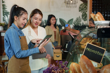Smiling Asian women in casual attire view tablet in coffee shop with prominent bread display,...