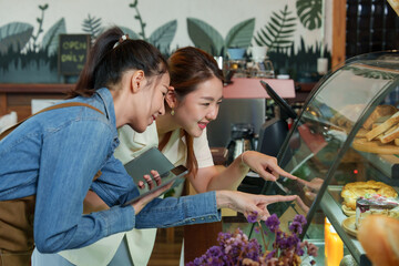 Young woman in blue apron, tablet in hand, consults with customer pointing at pastry display in a cozy, plant-adorned coffee shop.small family business