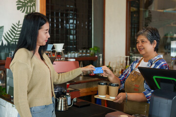 Smiling customer pay with blue credit card with barista at coffee shop, carrying tray with two takeaway cups, coffee equipment in backdrop. Cashless payments. small family business