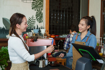 Inside a cozy coffee shop, Asian female barista in denim and apron accepting a credit card from...