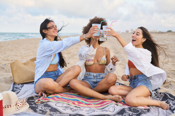 Three women are sitting on a blanket on the beach, holding up their drinks