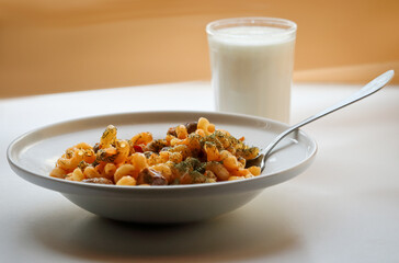 Spiral pasta in sauce with chicken, beans and vegetables covered with dill in a white plate on a kitchen table with metal fork and a glass of milk