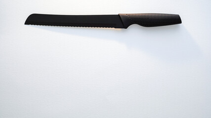 A black-handled serrated bread knife is placed on a clean white surface, illuminated by daylight,...