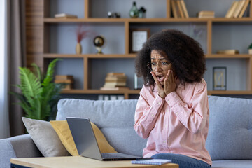 Shocked woman sitting on a couch at home, using a laptop and expressing astonishment. Surprised...