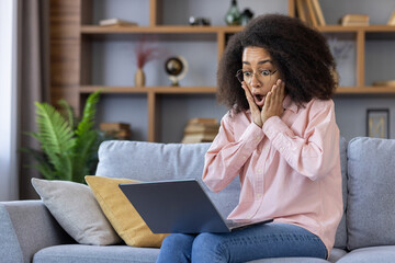 Woman in casual attire expressing shock and surprise while looking at her laptop screen at home