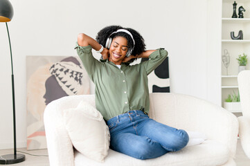 A cheerful African-American woman stretches with satisfaction on a comfortable sofa, wearing headphones, possibly taking a break from work or enjoying a leisurely moment in a cozy home environment