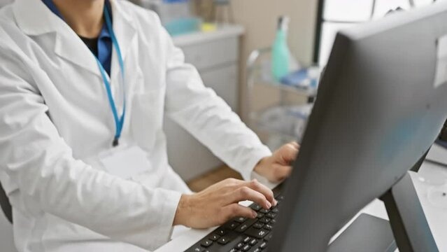 A focused hispanic man in a white lab coat working on a computer in a bright laboratory setting.