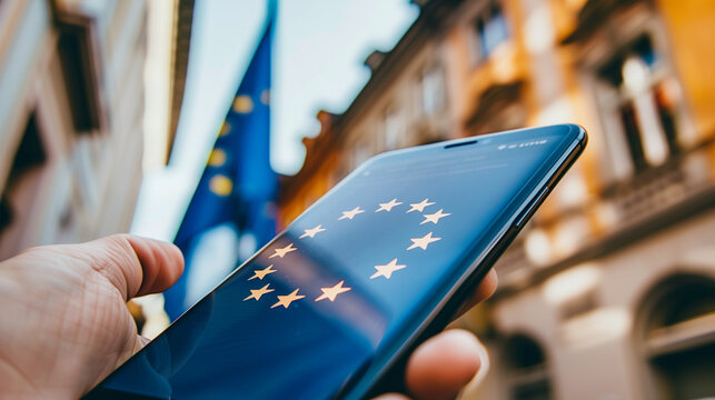 smartfon z ekranem wyświetlającym flagę Unii Europejskiej, leżący na pofałdowanej tkaninie r&oacute;wnież z motywem flagi UE. Jasne kolory flagi i kontrastujące tło tworzą wyrazisty obraz