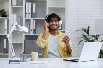 Happy man listening to music while working on a laptop from home. He is holding a smartphone and wearing headphones.