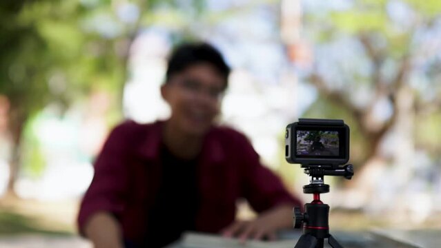 Asian man recording and talking to camera. Young man vlogging and taking selfie with action camera at public park.